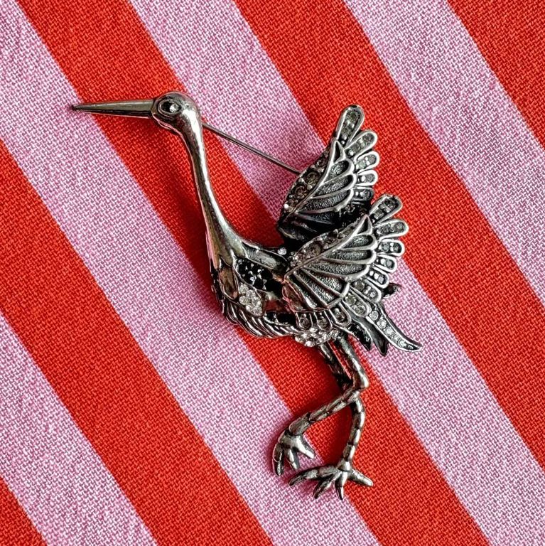 A silver crane pin on a pink and red background in a post about the one of a kind show.