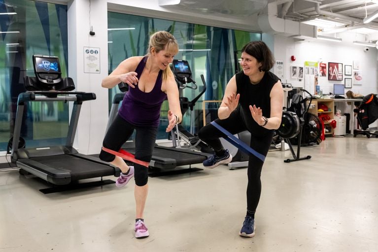Two women, Kelly and Gillian, smile while performing resistance band balance exercises side-by-side in a gym.