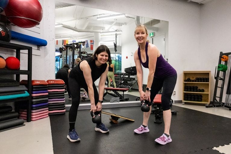 Kelly and Gillian smile at the camera while holding kettlebells in a bent-over stance during a gym workout.