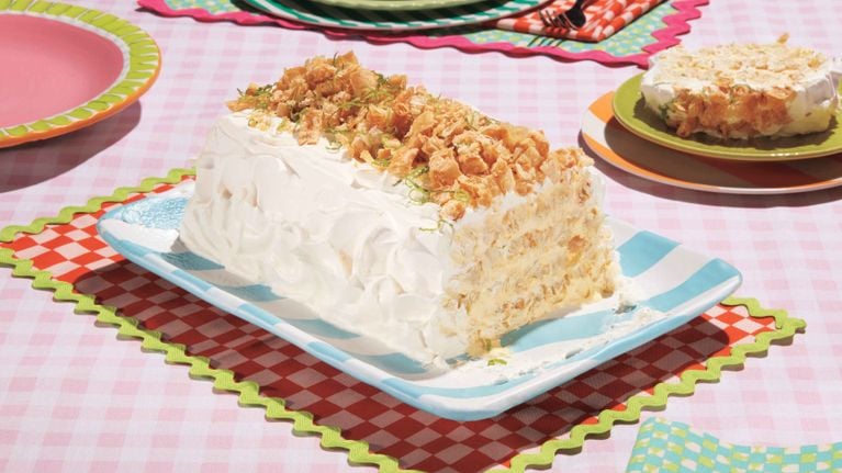 A rectangular Mango Lime Napoleon Cake is centered on a light blue and white striped platter. The cake is frosted with fluffy white whipped cream. The top is generously covered with golden, crunchy flakes and sprinkled with lime zest. The side facing the camera reveals the many layers of pastry and creamy filling. The platter rests on a red and white checkered placemat with a bright green scalloped edge. A second, smaller, round frosted cake is visible on a green plate in the upper right, and a pink plate is to the upper left, all on a pink and white checkered tablecloth.