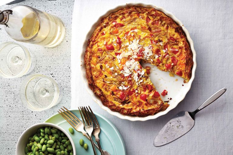 A gluten-free quiche with a golden-brown, shredded potato crust is presented in a white ceramic baking dish, with one slice already removed, revealing the savory, colorful filling. The filling appears to be a mix of eggs, melted cheese, and visible pieces of red pepper and onion. The quiche is topped with a sprinkle of shredded white cheese and is centrally placed on a light-colored, textured tablecloth. To the left, a teal-blue plate holds a small green bowl of edamame (or similar green peas), next to two silver forks. Above this, two clear glasses and a clear glass bottle containing a pale liquid (possibly white wine) are visible. A silver pie server/spatula rests on the right side of the quiche. The lighting is bright, highlighting the texture and color of the food.