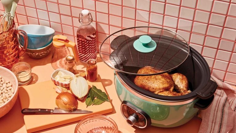 A slow cooker with a glass lid contains a whole, seasoned roast chicken resting on a wire rack. In the foreground, a cutting board holds ingredients for the recipe, including a halved onion, several cloves of garlic, bay leaves, and a small pile of dried white beans. Various spices in small jars, a large blue and white mixing bowl, and other kitchen items are visible on the counter in the background, set against a pink-tiled wall. The scene is brightly lit and focuses on the preparation for a slow-cooker meal.