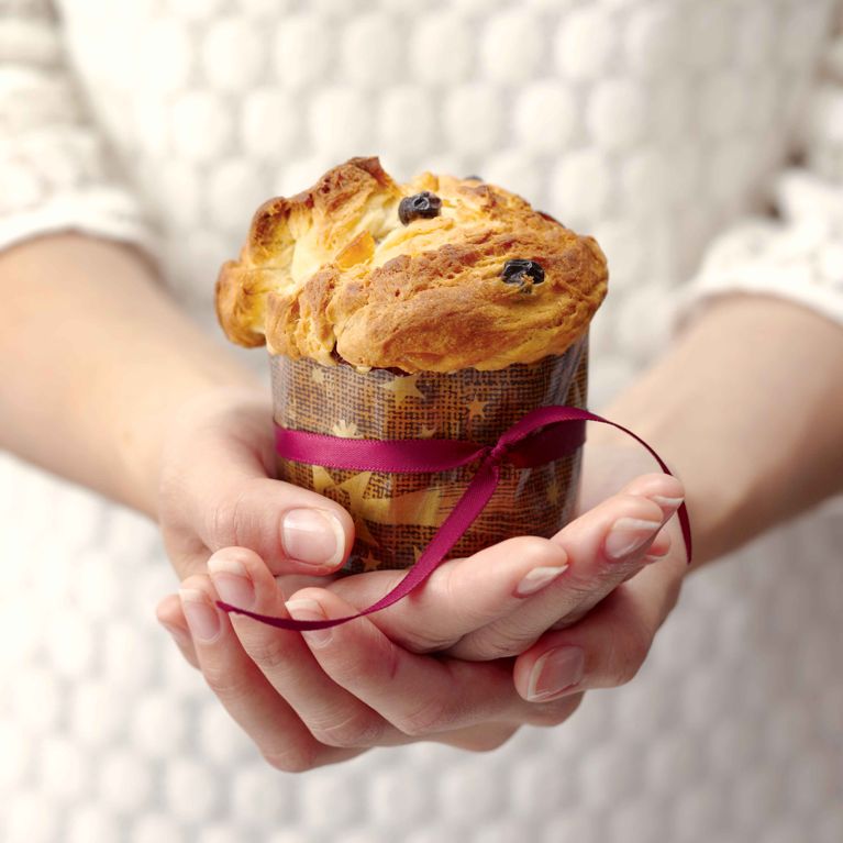 Alt text writer Custom Gem A close-up, square-cropped photograph focuses on two hands gently cupping a small, individual Panettone. The hands have neatly manicured nails and belong to a person wearing a white, textured knit sweater that is slightly out of focus in the background. The Panettone has a tall, brown paper baking cup wrapper, tied around the middle with a deep magenta or wine-colored ribbon, knotted with tails trailing down. The top of the cake is domed, golden-brown, and has a cracked surface, with visible dark raisins and candied fruit pieces baked into the rich, light yellow dough.