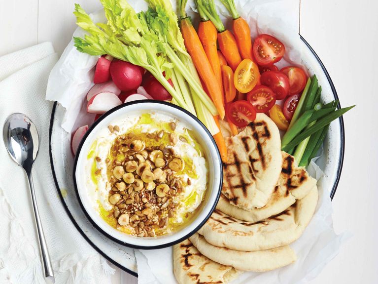 A bright overhead shot of a snack board on a white enamel tray. A small bowl of white labneh sits in the center, garnished with golden olive oil, hazelnuts, and seeds. It is encircled by colorful dipping items: celery sticks, whole carrots with greens, red and yellow cherry tomatoes, scallions, radishes, and toasted pita bread with grill marks. A silver spoon rests on a napkin to the left.