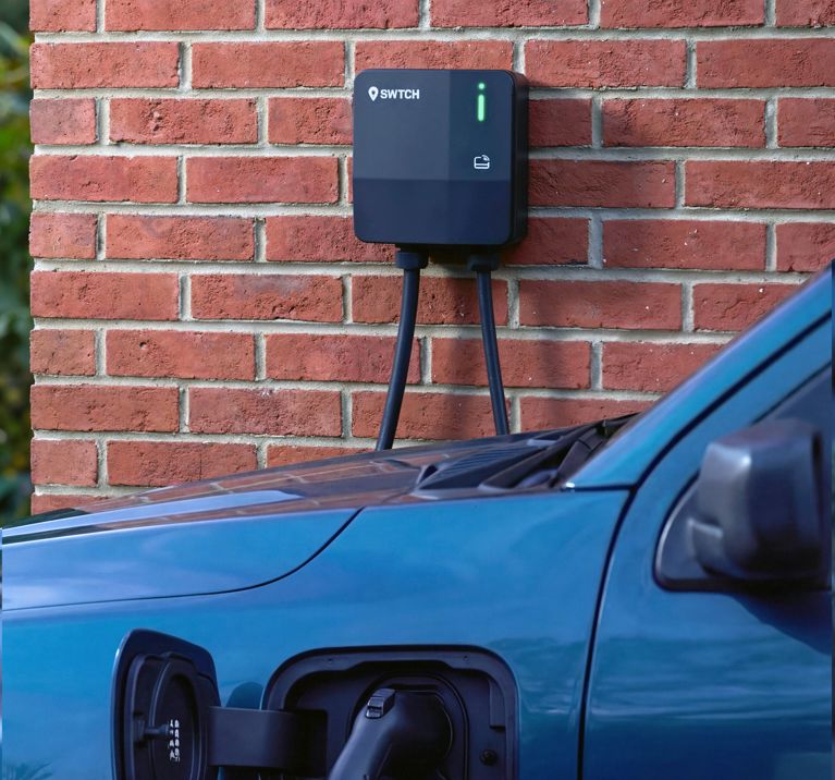 A black SWTCH electric vehicle charger is mounted on a red brick wall. The charger’s green indicator light is on, showing it is active. In the foreground, the charging cable is plugged into the side port of a blue car, with the charging flap open.