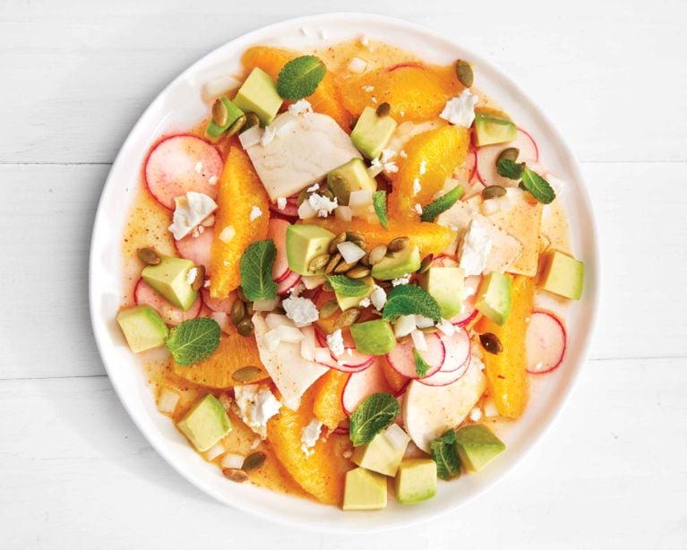An overhead shot of a vibrant, colorful salad served on a white round plate against a white wooden table. The salad features juicy orange segments, cubes of fresh green avocado, thin rounds of red radish, and slices of white jicama. It is garnished with crumbled white cheese, toasted pumpkin seeds, and fresh mint leaves.