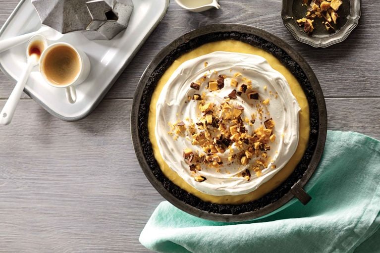 An overhead shot of a whole banana caramel cream pie sitting on a grey wood surface. The pie features a dark chocolate cookie crumb crust encircling a ring of yellow custard. The center is generously topped with a swirl of white whipped cream and sprinkled with chopped pieces of golden, chocolate-covered honeycomb toffee. To the left, a white serving tray holds a moka pot and a small cup of espresso. A teal cloth napkin rests beneath the pie pan on the right.