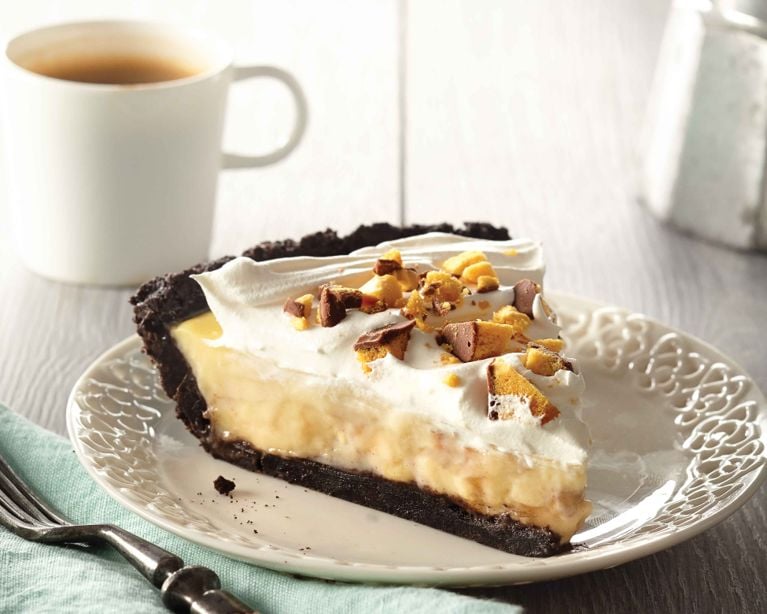 An overhead shot of a whole banana caramel cream pie sitting on a grey wood surface. The pie features a dark chocolate cookie crumb crust encircling a ring of yellow custard. The center is generously topped with a swirl of white whipped cream and sprinkled with chopped pieces of golden, chocolate-covered honeycomb toffee. To the left, a white serving tray holds a moka pot and a small cup of espresso. A teal cloth napkin rests beneath the pie pan on the right.