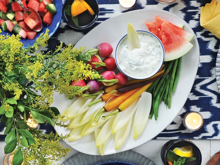 An overhead shot of a vibrant crudité platter featuring a bowl of creamy leek dip with green speckles. An endive leaf rests in the dip, which is surrounded by fresh dippers including red radishes, rainbow carrots, green beans, Belgian endive leaves, and watermelon slices. The setting includes yellow wildflowers, a side of watermelon salad, cocktails, and votive candles on a blue and white patterned table runner.