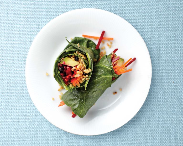 Top-down view of two healthy vegetable wraps served on a white plate against a light blue, textured background. Large, dark green collard leaves serve as the wrap, holding a vibrant filling of golden curried quinoa, shredded orange carrots, julienned red beets, avocado slices, and dark dried fruit. One wrap lies horizontally while the other rests on top with the cut end facing up, showcasing the colorful ingredients.