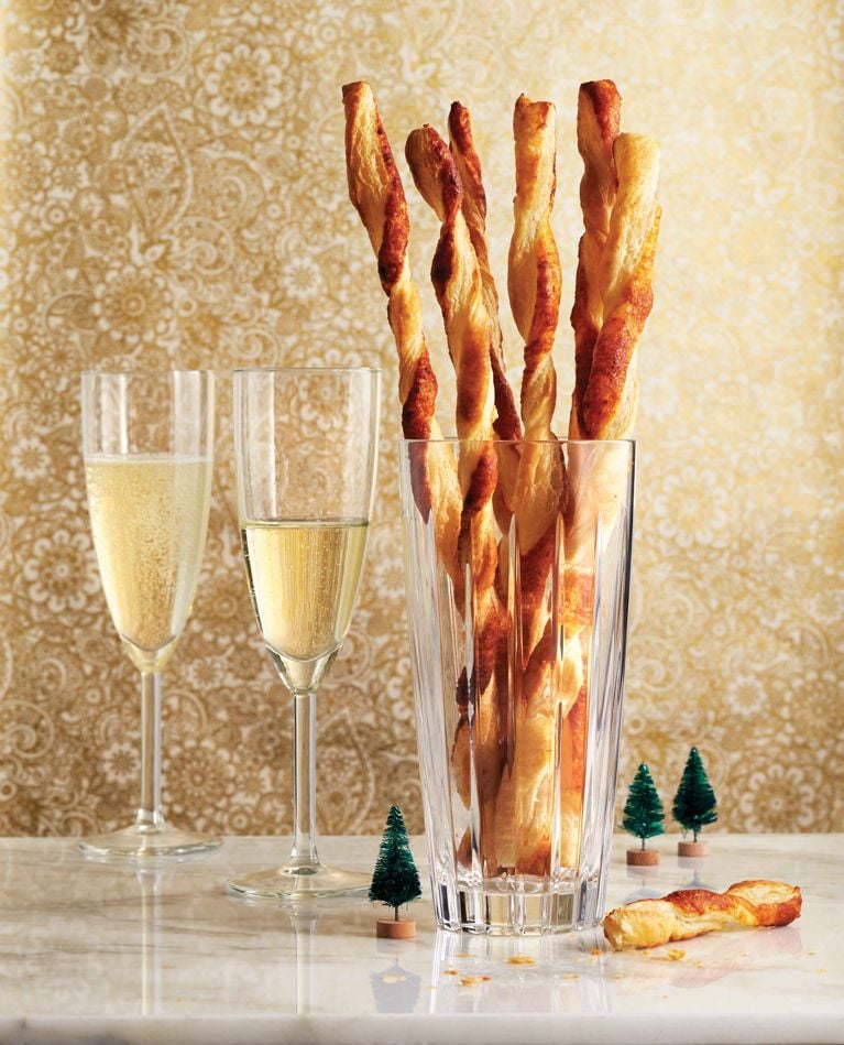 A festive holiday arrangement featuring a tall, cut-crystal glass filled with long, twisted puff pastry cheese straws standing vertically. To the left, two champagne flutes are filled with bubbly sparkling wine. The scene is set on a white marble surface against an elegant gold-and-white damask wallpaper background, decorated with three tiny green bottle-brush Christmas trees scattered near the base of the glass.