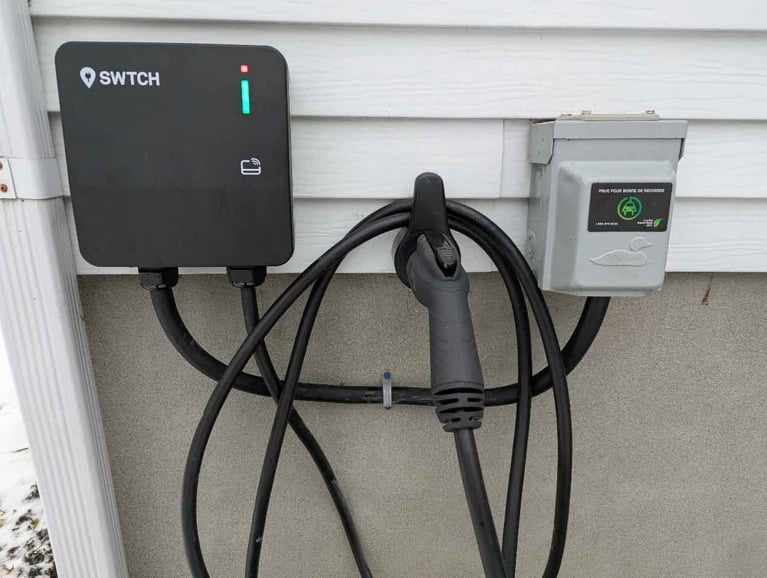A close-up of a black SWTCH brand electric vehicle charger mounted on a white-sided exterior wall. A green status light is illuminated on the front panel. The charging cable is neatly coiled on a wall hook, with the charging nozzle resting in the center. To the right is a grey electrical disconnect box with a green leaf logo.