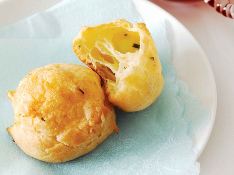 Two golden-brown Gruyère cheese puffs rest on a light blue paper napkin. One puff is whole, displaying a crisp, rounded exterior, while the other is broken open to reveal a hollow, airy, yellow interior speckled with herbs.