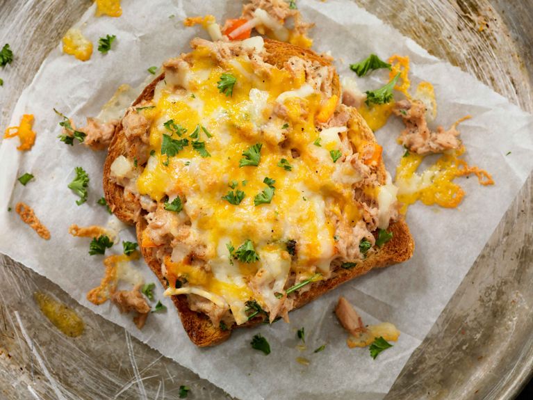 A close-up, top-down view of an open-faced spring tuna melt served on a slice of golden-brown toasted bread. The tuna salad is generously topped with melted yellow and white cheese and garnished with chopped fresh parsley. The toast sits on crinkled white parchment paper atop a rustic, scratched metal tray, surrounded by scattered bits of crispy cheese and herbs.