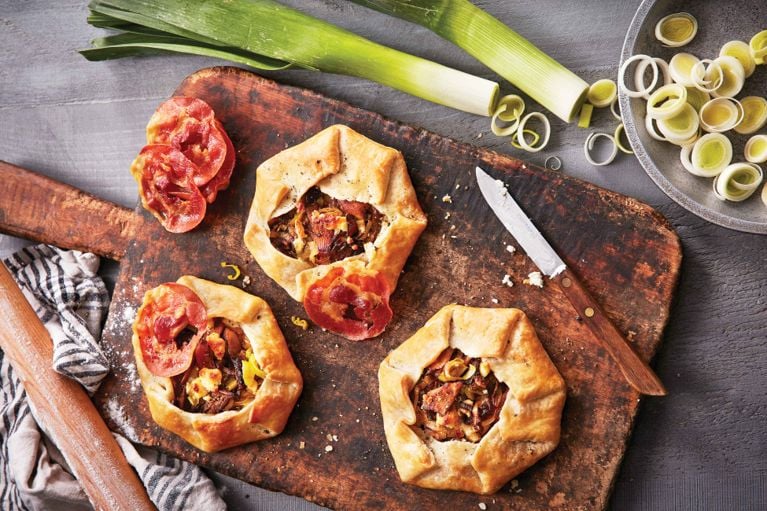 An overhead flat-lay photograph showing three rustic, single-serving savory galettes arranged on a dark, worn wooden cutting board. The galettes feature golden-brown pastry crusts folded roughly over a filling of caramelized leeks and cheese. One galette is topped with a crispy round of pancetta or prosciutto, and a separate pile of these cooked meat slices sits on the upper left of the board. The scene includes fresh raw leek stalks at the top, a bowl of sliced raw leek rings to the right, a small paring knife, and a flour-dusted rolling pin resting on a striped kitchen towel to the left.