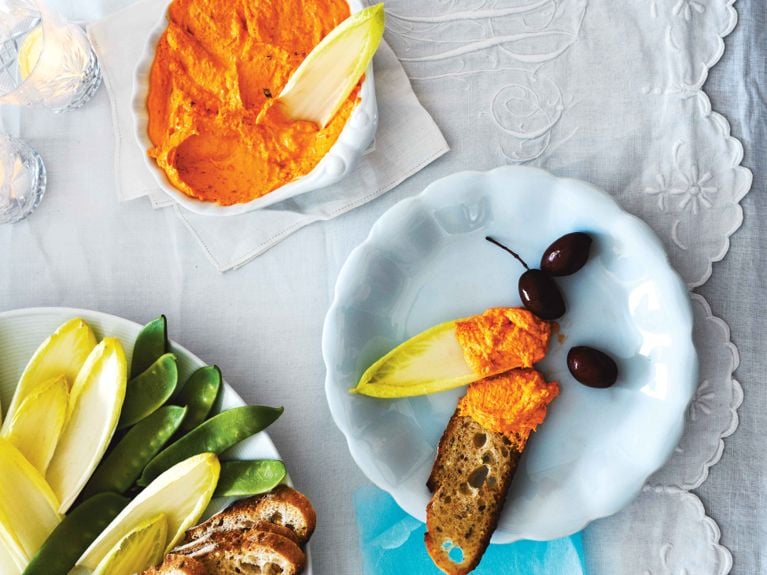 An overhead view of a bright orange molten goat cheese dip served in a white scalloped bowl, with an endive leaf dipped into the mixture. To the left, a platter holds fresh snap peas, Belgian endive leaves, and slices of toasted baguette. On the right, a small blue plate displays a serving of the dip spread on a slice of toast and an endive leaf, accompanied by black olives, all set against a white embroidered tablecloth with votive candles nearby.