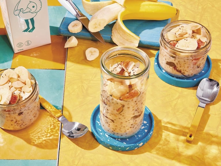 Overnight chia oatmeal in three different mason jars—two short and one tall—on a yellow table beside a sliced banana and yellow spoons and a carton of oat milk