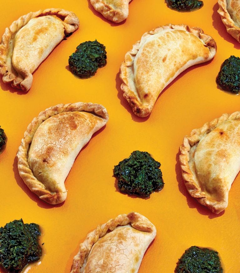 Golden-brown potato and chorizo empanadas served with chimichurri sauce on a colorful orange background.