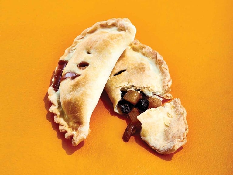 Two baked, crescent-shaped empanadas rest on a bright orange surface. The top empanada has a golden-brown crimped crust with dark syrup bubbling out of small vents. The second empanada is broken open, revealing a chunk of the flaky crust and a filling of diced potatoes and dark raisins.