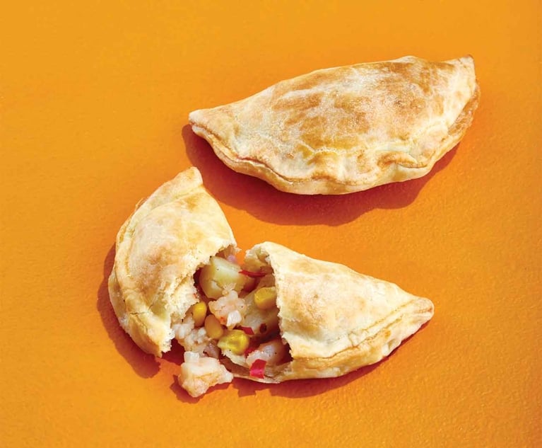 Two golden-brown baked empanadas sit on a bright orange background. The top empanada is whole, showing a flaky, sealed crescent edge. The bottom empanada is split open in the center, revealing a chunky filling of chopped shrimp, yellow corn kernels, diced potatoes, and specks of red pepper.