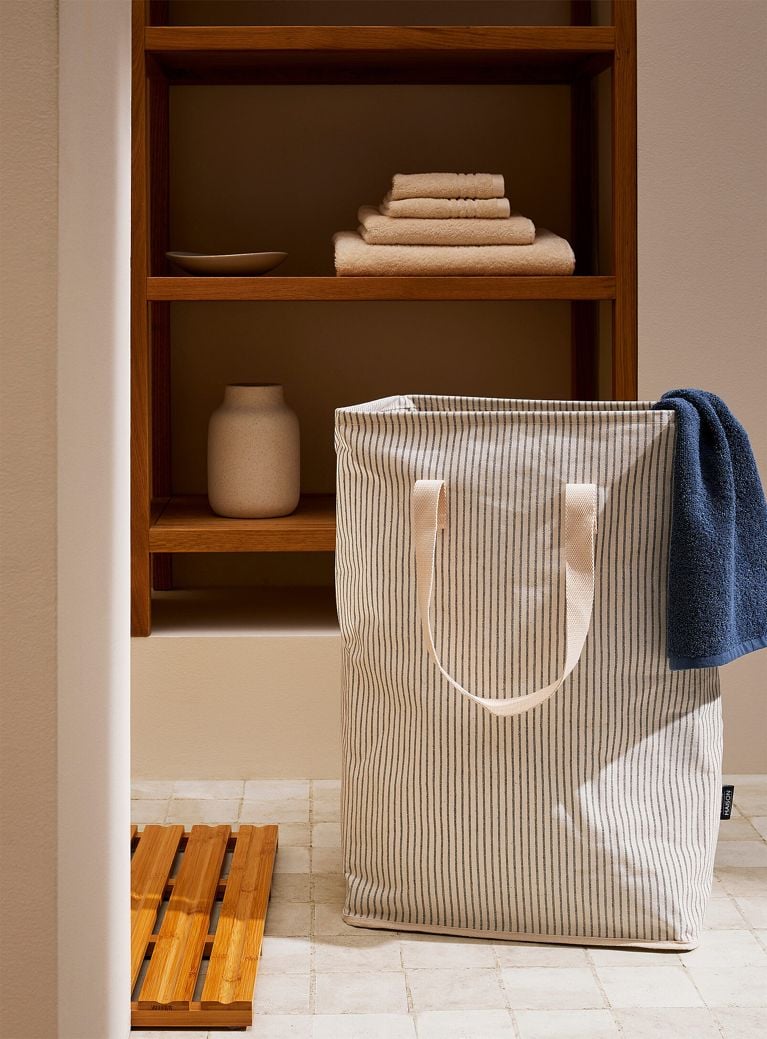 A tall, rectangular fabric laundry hamper featuring a pattern of thin, vertical dark charcoal stripes on a cream background. The hamper has long, cream-colored webbed handles, and a dark blue towel is draped casually over the right rim. It sits on a light, square-tiled floor next to a wooden slat bath mat. In the background, a built-in wooden shelving unit holds a neat stack of folded beige towels, a small dish, and a speckled white ceramic vase.