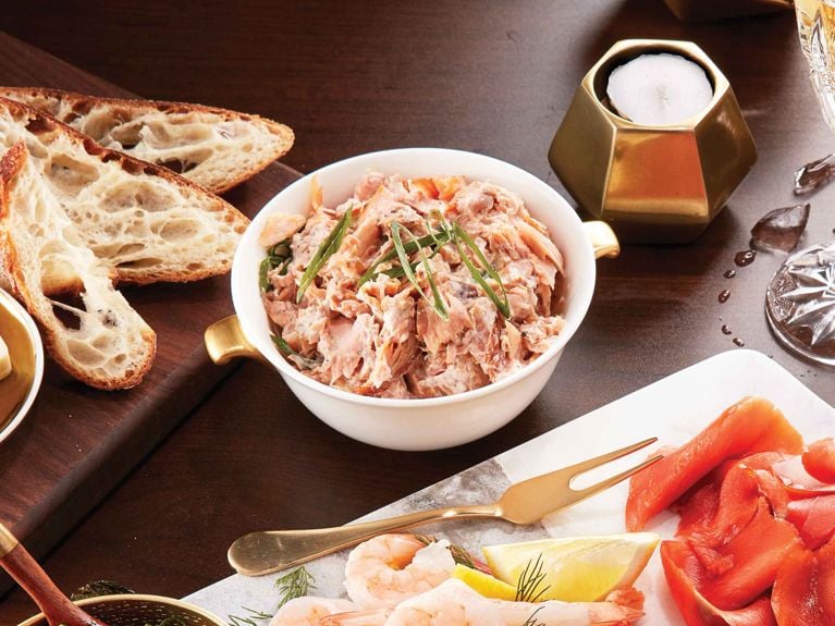 A white ceramic bowl filled with creamy, chunky smoked trout dip garnished with thin strips of green onion. To the left, slices of toasted rustic bread rest on a dark wooden board. The dip is part of an elegant appetizer spread, with a marble platter in the foreground holding shrimp, lemon wedges, and smoked salmon, accompanied by a gold serving fork and a geometric gold candle holder in the background.