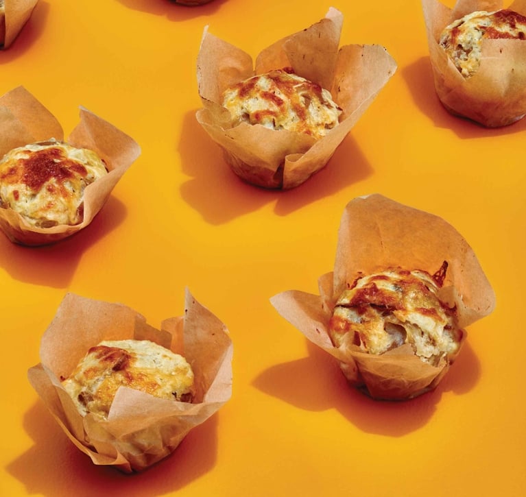 A high-angle, patterned shot of savoury smoky Gouda muffins arranged on a solid, vibrant orange background. Each muffin is baked in a rustic, unbleached parchment paper tulip liner. The tops are golden-brown and textured with melted, toasted cheese. Hard studio lighting casts distinct, dark shadows to the bottom right of each muffin.
