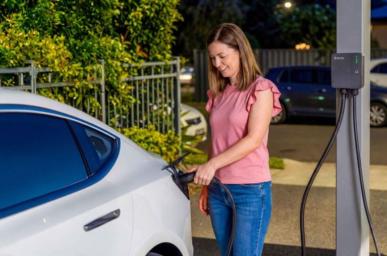 A close-up of a black SWTCH brand electric vehicle charger mounted on a white-sided exterior wall. A green status light is illuminated on the front panel. The charging cable is neatly coiled on a wall hook, with the charging nozzle resting in the center. To the right is a grey electrical disconnect box with a green leaf logo.