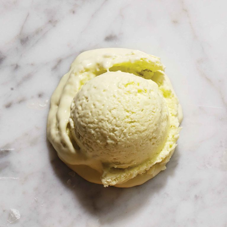 A top-down view of a single scoop of pale, pastel green-yellow avocado ice cream resting directly on a white marble surface. The scoop has a textured, creamy surface and is melting slightly, forming a thick, liquid pool around the base.