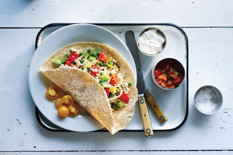 Alt text writer Custom Gem An overhead shot of a tofu scramble wrap served on a white metal tray against a white painted wooden background. The wrap, made with a spotted tortilla, is folded over a filling of crumbled tofu, diced red peppers, green peas, and chopped green onions. Several halved yellow cherry tomatoes rest on the plate beside the wrap. To the right, two small bowls contain a white cream and a red salsa, alongside a tiny bowl of salt. A vintage knife and fork with yellowed handles lie next to the plate.
