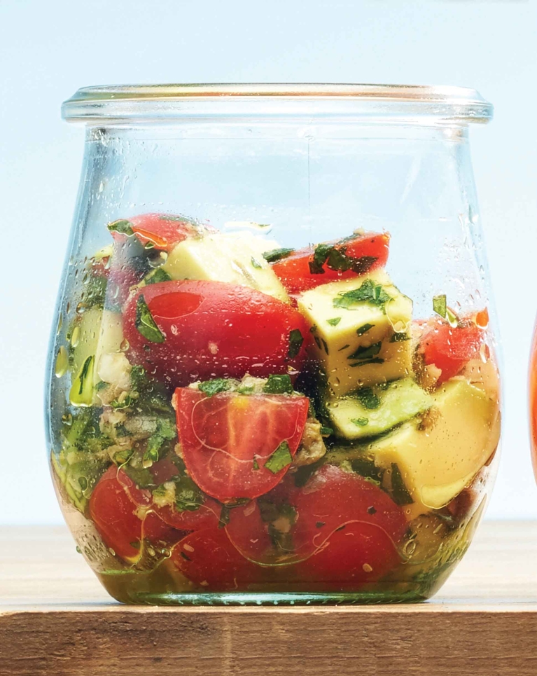 A close-up of a clear glass jar filled with chunky tomato and avocado salsa. The mixture contains halved red cherry tomatoes, anchovies, cubed green avocado, and chopped fresh herbs tossed in a light dressing, sitting on a wooden surface.