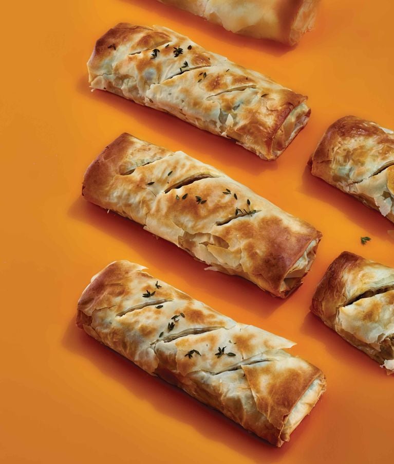 Baked turkey and mushroom strudels wrapped in golden, flaky phyllo pastry. The rolls are scored diagonally, garnished with fresh thyme, and set against a vibrant orange surface.