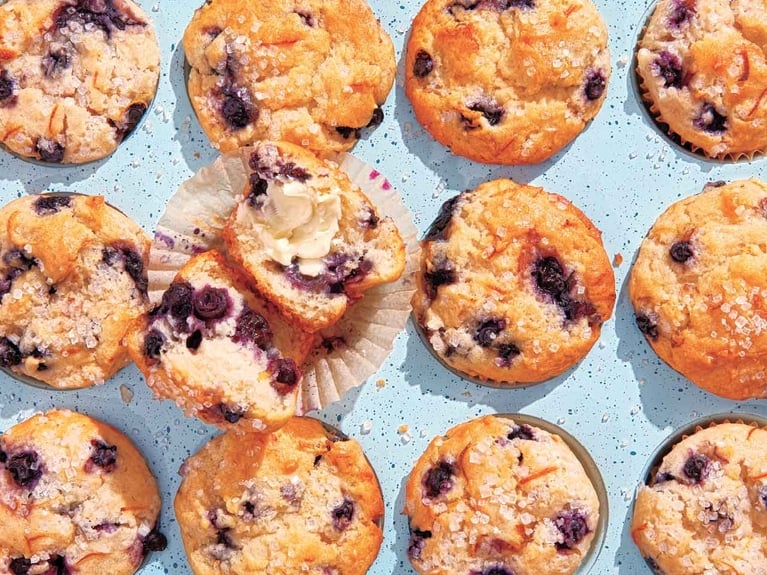 An overhead shot of a light blue, speckled muffin tin filled with golden-brown blueberry muffins. Coarse sugar crystals are sprinkled over the domed tops, which are dotted with dark berries. One muffin in the center-left is split open, revealing a fluffy interior topped with a dollop of melting butter.
