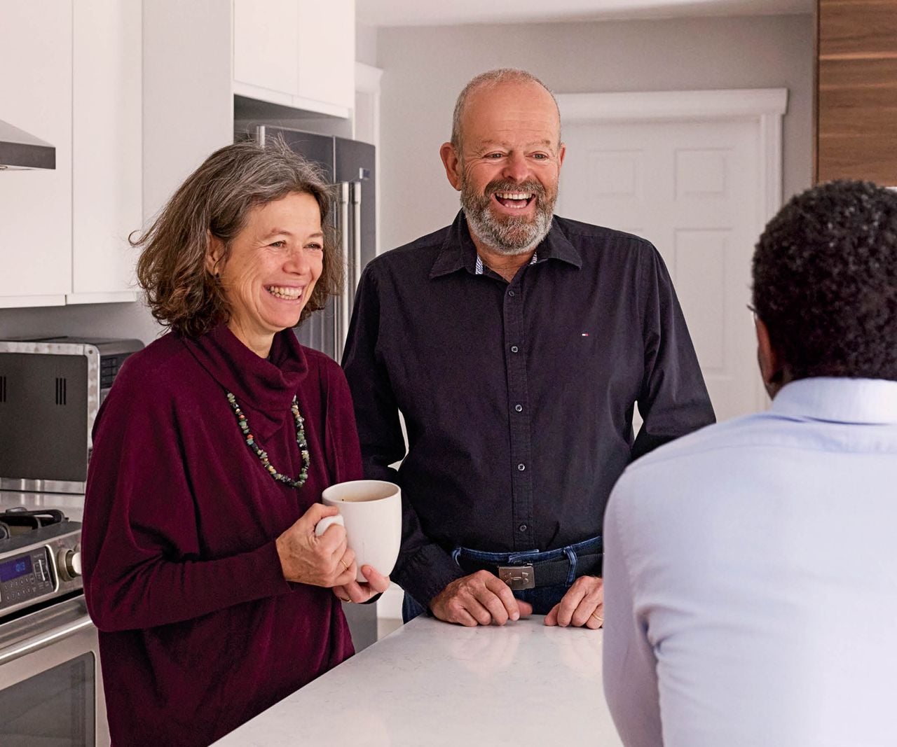 A man and woman stand at a kitchen counter smiling with a man, whose back is to the camera. Raphaëlle Curis and Michel Racine found common ground with John over shared traditions and food.