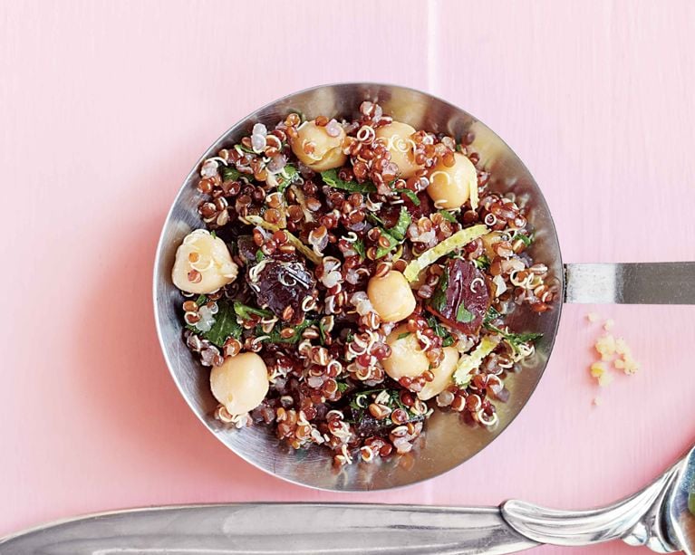 A vibrant grain salad served in a round stainless steel measuring cup. The base consists of fluffy red quinoa mixed with plump, light-tan chickpeas and chunks of dark, mahogany-colored dates. Specks of bright green mint or parsley and thin yellow lemon zest are scattered throughout. The dish is photographed from directly above on a smooth pink surface, with the edge of a vintage silver spoon visible at the bottom.