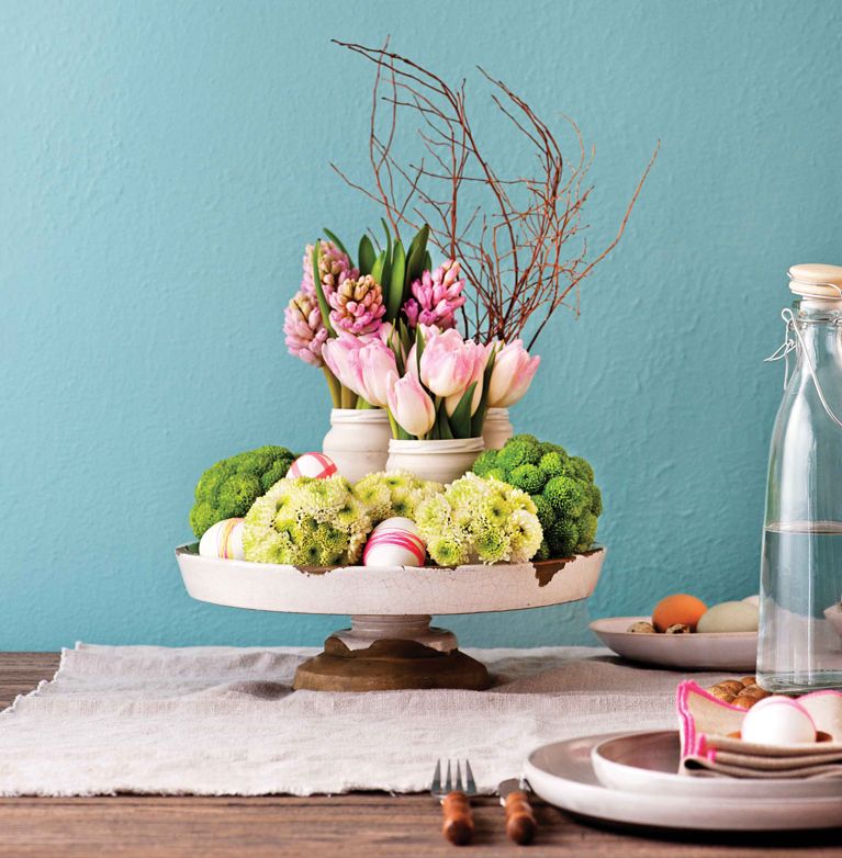 A tiered Easter centerpiece on a white pedestal stand featuring pink tulips, hyacinths, and thin brown branches. The base is decorated with green moss-like flowers and white eggs wrapped in pink twine, set against a bright teal wall.