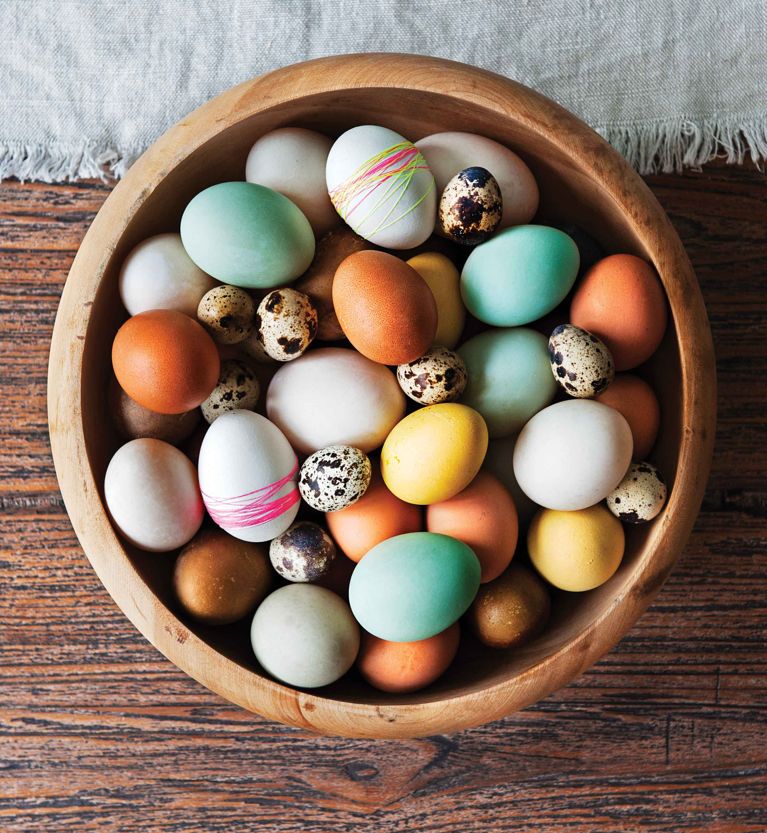 A wooden bowl overflows with a curated mix of eggs, showcasing a palette of spring colors. The collection includes smooth chicken eggs dyed in soft teal, pale yellow, and earthy orange, alongside natural white and brown eggs. Several white eggs are wrapped in thin pink and green neon thread. Interspersed throughout are small, heavily speckled quail eggs. The bowl sits on a dark, grained wooden surface partially covered by a light grey linen cloth.