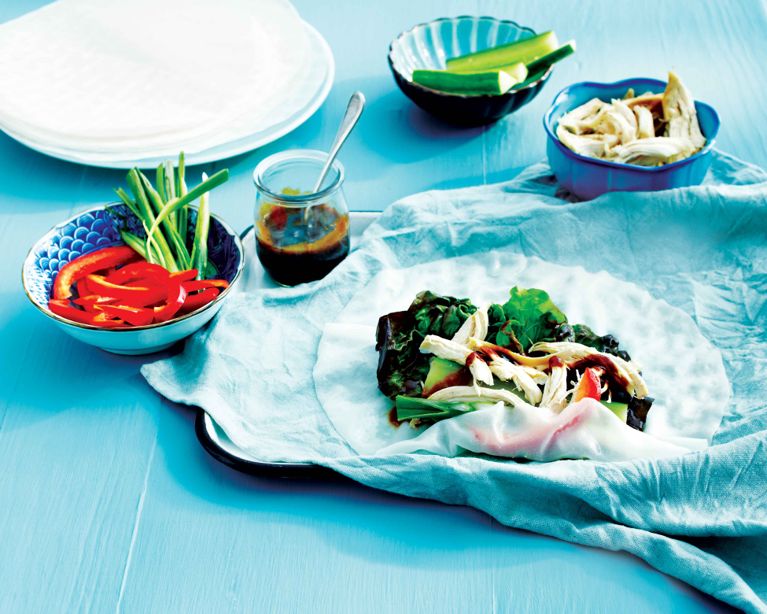 High-angle shot of a DIY meal station on a blue surface. In the center, a translucent rice paper wrap rests on a crumpled light blue napkin, filled with dark leafy greens, shredded chicken, and a drizzle of dark hoisin sauce. Surrounding the wrap are small blue bowls containing sliced red bell peppers, green scallions, cucumber spears, and extra chicken. A small glass jar of hoisin sauce with a spoon sits nearby