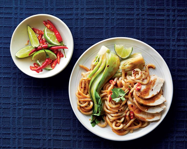 Overhead view of a white plate containing thick udon noodles coated in a savory brown sauce. The dish is topped with sliced chicken breast, steamed bok choy, and a sprig of cilantro. To the left is a small bowl filled with red bird's eye chilies and lime slices, all set against a textured navy blue tablecloth.