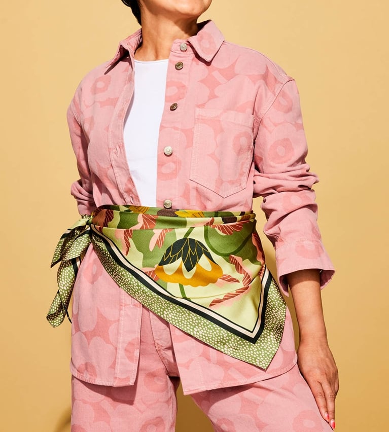 A model wearing a green silk scarf tied around her waist over a pink floral denim jacket on a yellow background.