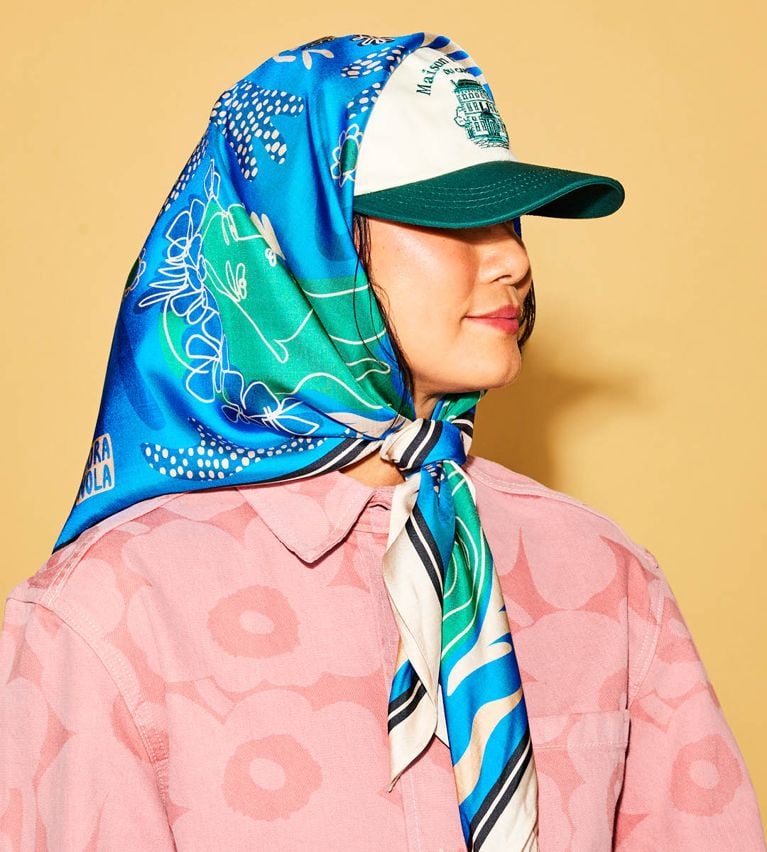 A model wearing a blue silk scarf tied around her head over a green baseball hat on a yellow background.