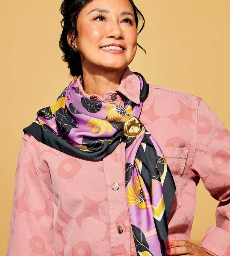 A model wearing a pink and purple silk scarf with a brooch over a pink floral denim jacket on a yellow background.