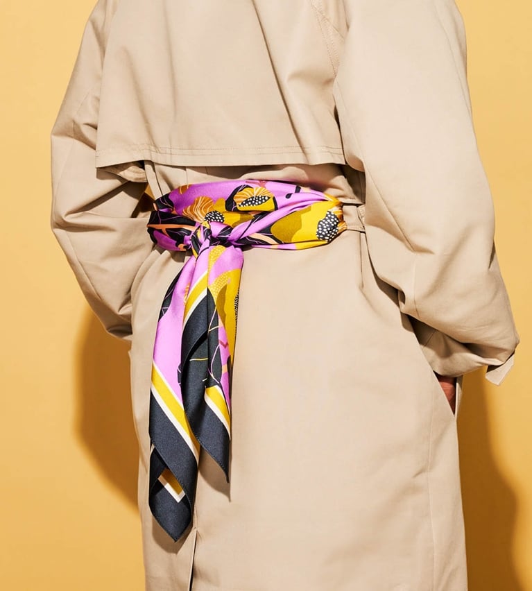 A model wearing a purple and yellow silk scarf tied around the loops of a trench coat on a yellow background.
