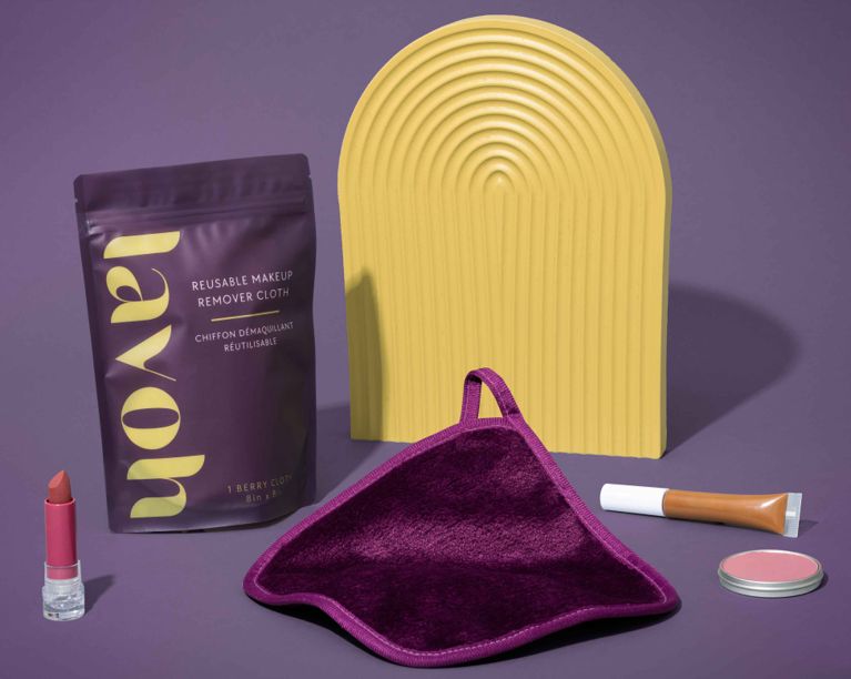 A flat-lay product photography shot against a deep purple background featuring a Lavoh Reusable Makeup Remover kit. The set includes a dark berry-colored microfiber cloth with a hanging loop, its matching purple resealable packaging with yellow "lavoh" branding, and a yellow arched decorative prop. Surrounding the items are makeup products including a pink lipstick, a concealer tube, and a small tin of blush.