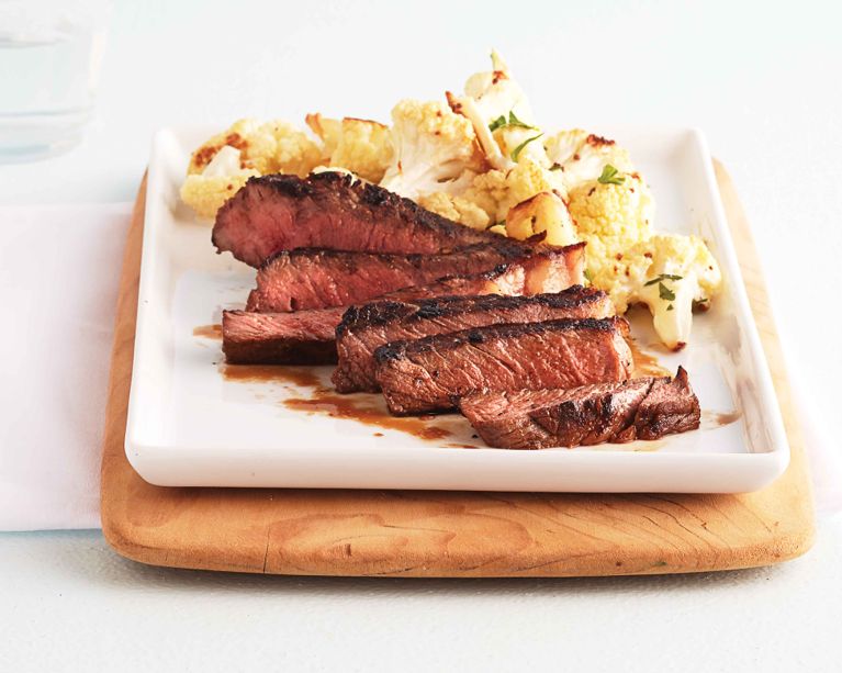A white plate features several thick slices of seared steak, showing a pink medium-rare center and a dark brown crust. In the background, golden-brown roasted cauliflower is garnished with fresh herbs. The plate sits on a wooden board against a clean, bright white background.