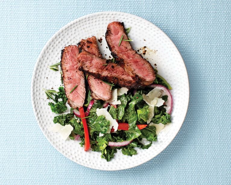 Top-down view of a gourmet meal: several thick slices of seared steak showing a pink, medium-rare center and a dark charred crust. The steak sits on a vibrant green kale sauté mixed with slivers of red onion and red bell pepper. The dish is finished with large shavings of parmesan cheese and scattered rosemary sprigs, presented on a white dotted plate against a light blue textured background.