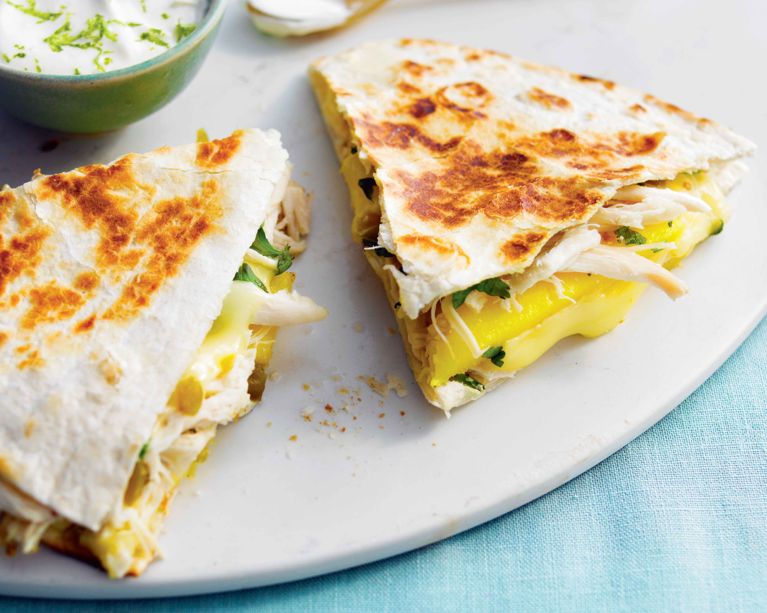 A close-up shot of two sliced mango and chicken quesadillas on a white plate. The flour tortillas feature charred, golden spots from toasting. The visible filling includes thick, vibrant yellow mango slices, melted creamy brie, shredded white meat chicken, and fresh green cilantro. In the upper left corner, a small teal bowl contains a white dipping sauce garnished with fresh green lime zest.