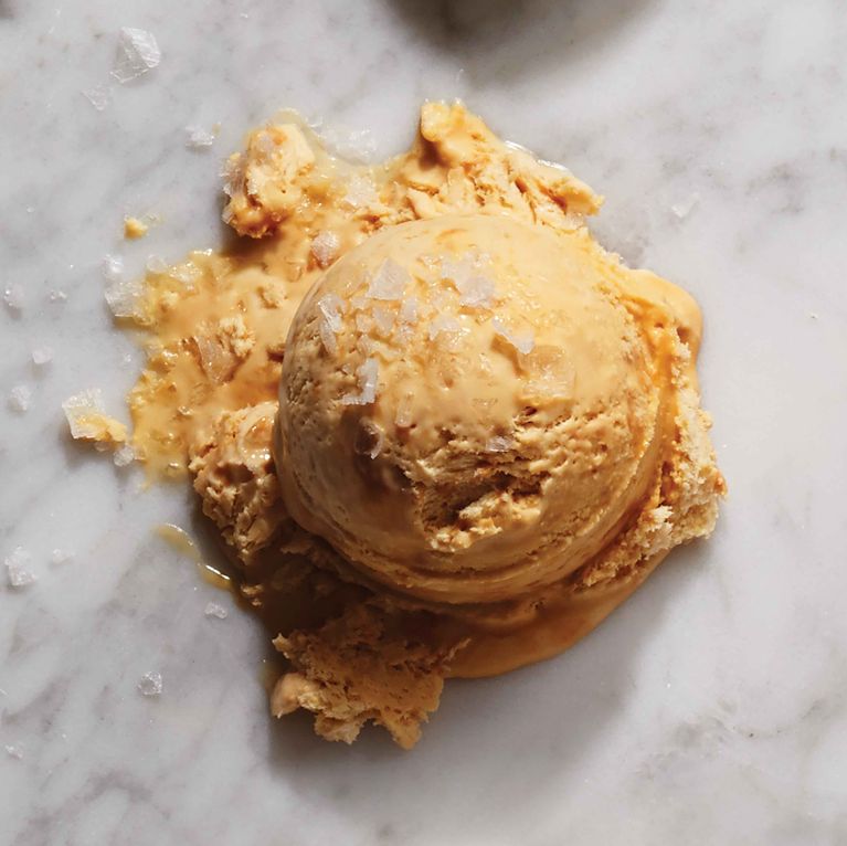 A single, large scoop of golden-tan salted caramel ice cream sitting on a white marble surface. The ice cream is slightly melting at the base and topped with large, translucent flakes of sea salt.