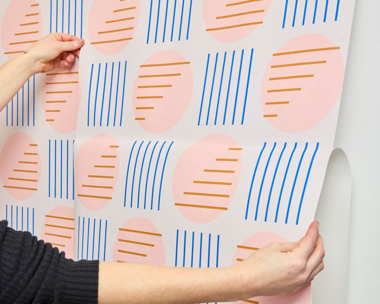 A woman installing peel-and-stick wallpaper aligns the pattern of a second panel on a white wall.