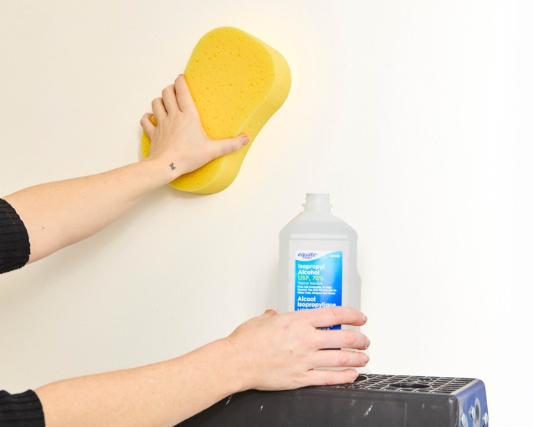 A woman cleans a wall with isopropyl alcohol and a sponge to prepare for installing peel-and-stick-wallpaper.